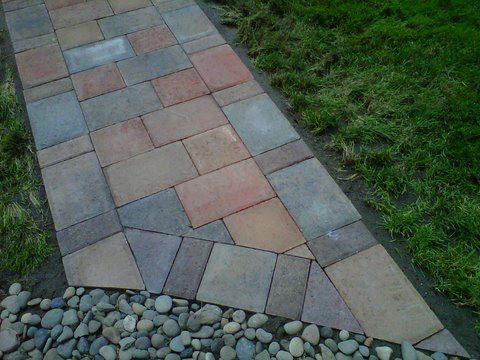 Paver pathway leading to gravel. Made with multiple shades of red, brown, and grey. Mowed lawn on either side.