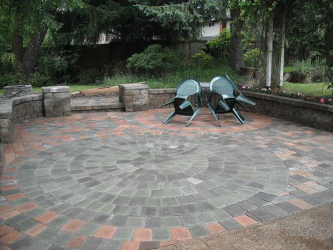 Rounded paver patio shape. Made with different shades of red, brown, and grey pavers. Small table and two chair to the right. Wall encompassing the patio.