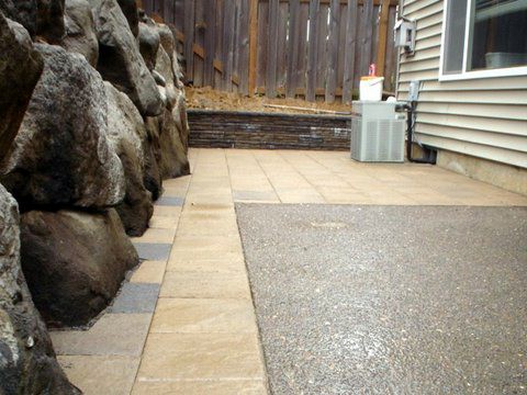 On the left is a retaining wall made out of natural rocks. Wooden fence in the back. Floor between house and wall made out of tan and grey pavers.