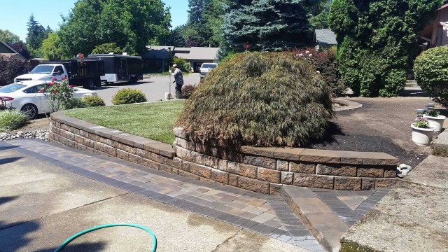 Retaining wall made out of brick pavers or natural stone with pathway and small trees trimmed and lawn maintained