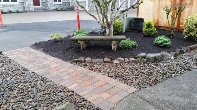 pathway made out of brick pavers and a natural stone bench with pathway and small trees trimmed and mulched soil maintained