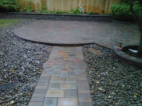 Rounded paver patio shape with pathway attached. Made with different shades of red, brown, and grey pavers. Surrounded by gravel. Wooden fence in the back.